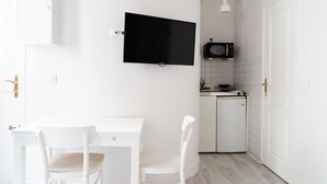 A small, white kitchen with a microwave, a stove, and a TV mounted on the wall. There is a white table with two chairs and a door with a golden handle.
