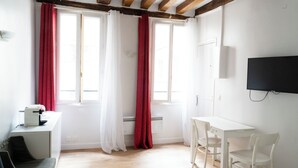 A room with a white desk, a chair, a television mounted on the wall, and a window with red curtains.