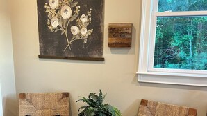 A room with two wooden chairs with woven backs, a small round wooden table with a plant, a wall-mounted painting of flowers, and a window with a view of trees. - The Butterfly Suite exudes an air of timeless elegance and natural splendor. Named in homage to the myriad species of butterflies that adorn the surrounding landscape. (Jasper)
