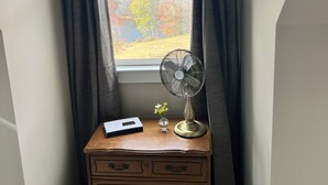 A corner room with a window, a wooden dresser, a fan, and a small vase with flowers. - The Butterfly Suite exudes an air of timeless elegance and natural splendor. Named in homage to the myriad species of butterflies that adorn the surrounding landscape. (Jasper)