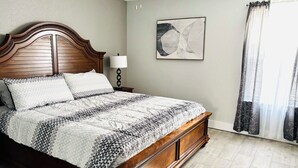 A bedroom with a wooden bed, a nightstand, a lamp, a ceiling fan, a framed abstract painting, and a window with curtains. - Home Sweet Home (Fayetteville)