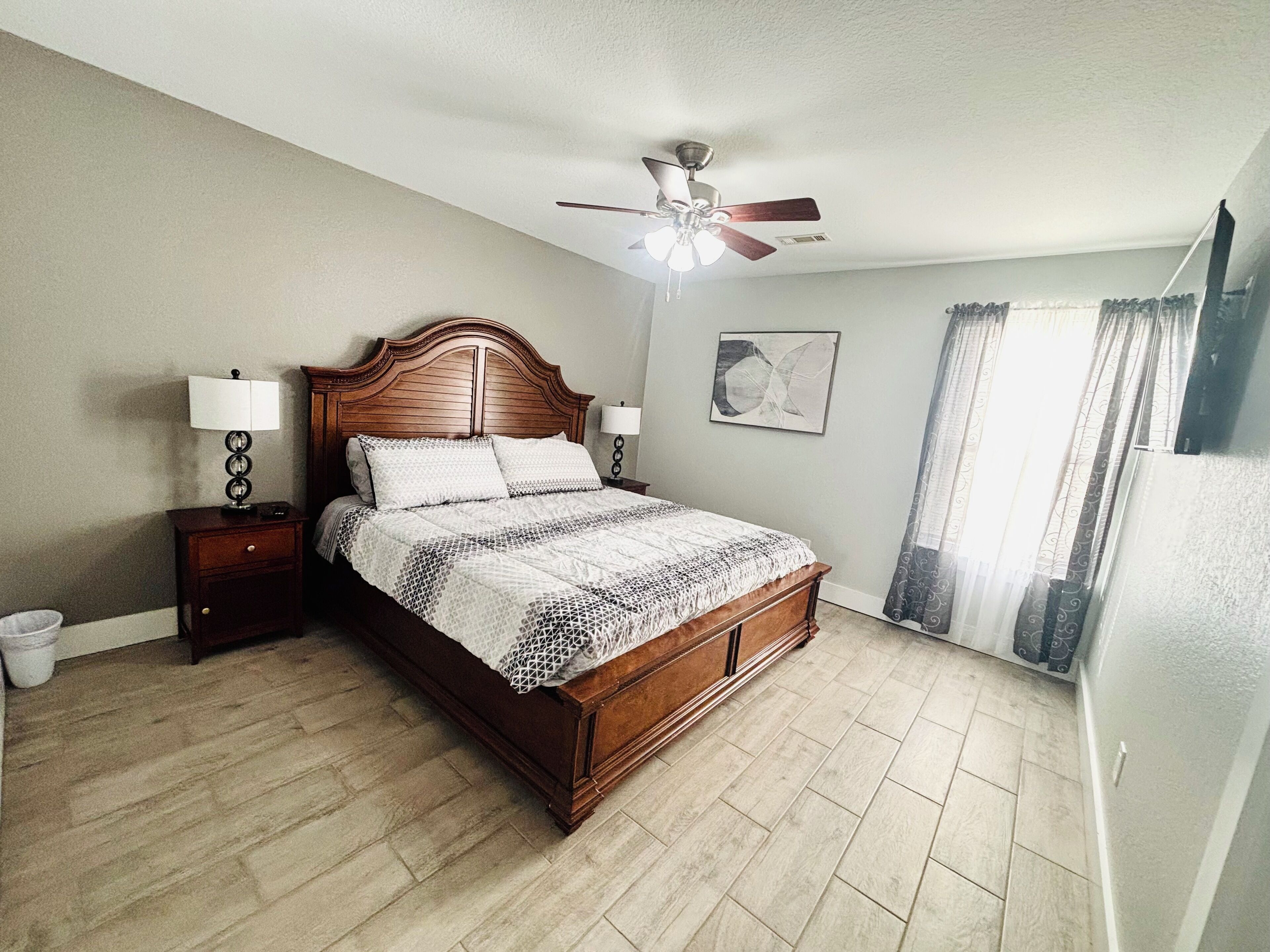 A bedroom with a wooden bed, two bedside tables with lamps, a ceiling fan, a large window with curtains, and a wall-mounted artwork.