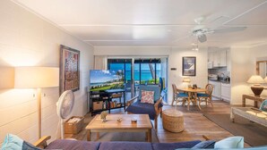 A living room with a sofa, a coffee table, and a TV. There is a view of the ocean through the glass doors.