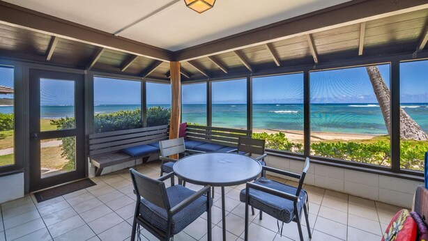 A covered outdoor seating area with a round table and chairs, large windows with a view of the beach and ocean, and a wooden ceiling.