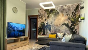 A modern living room with a flat-screen TV, grey sofa, and a decorative leaf mural on the wall.