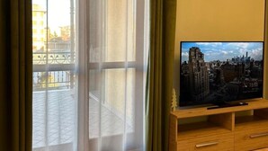 A living room with a large window, sheer curtains, a clock on the wall, and a flat-screen TV on a wooden stand.