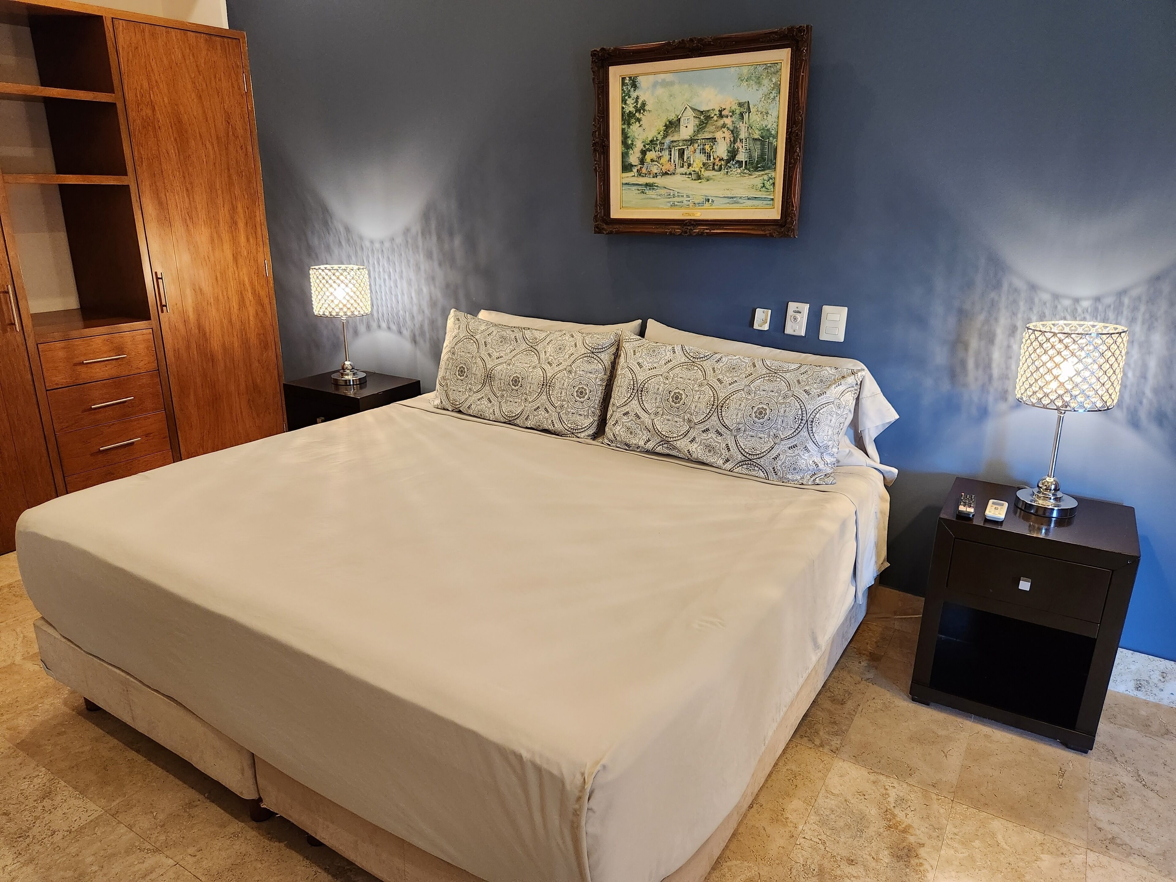 A bedroom with a large bed, two bedside tables with lamps, a wooden wardrobe, and a painting on the wall.