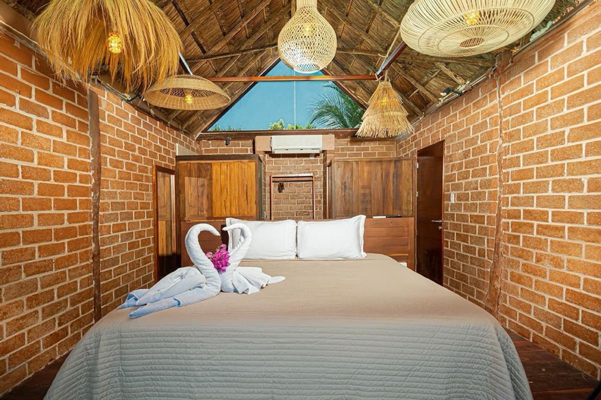 Habitación de hotel con pared de ladrillo, puerta de madera, cama con colcha blanca y dos lámparas tejidas.