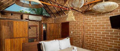 Un dormitorio con pared de ladrillo, muebles de madera, cama con ropa blanca y techo de paja con lámparas colgantes.