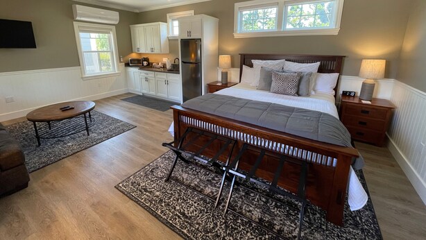 A bedroom with a bed, bedside tables, a nightstand, a ceiling fan, a kitchen area with cabinets and appliances, and a window.