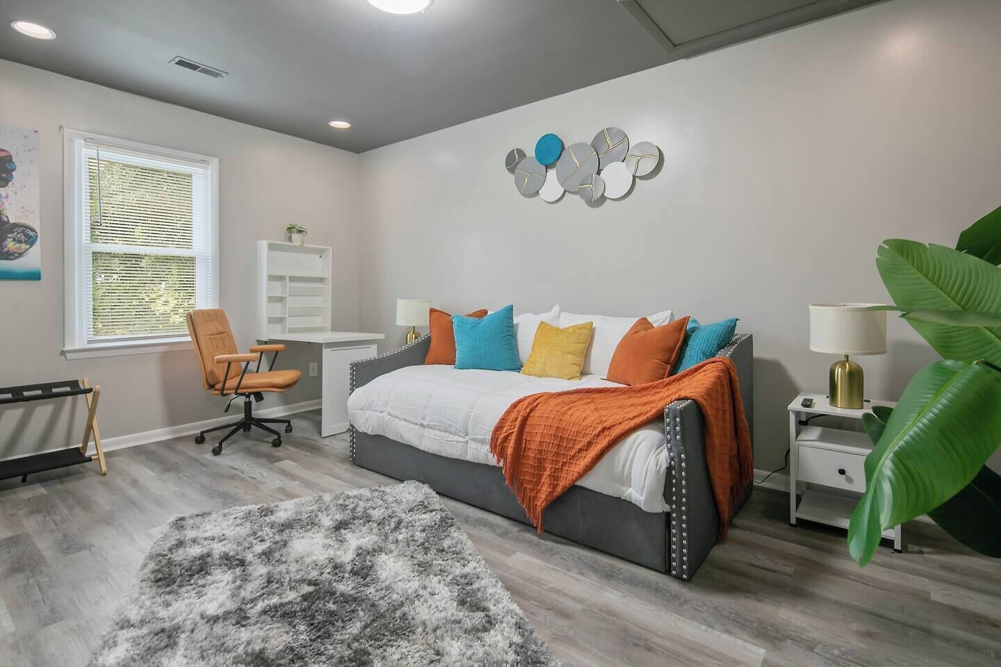 A bedroom with a bed, a desk, a chair, a nightstand, and a large plant.