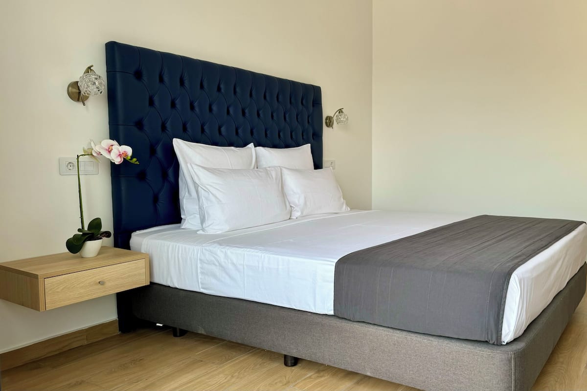 Una cama bien hecha con sábanas blancas y una manta gris, un cabecero capitonado azul oscuro y una mesita de noche con una planta y una lámpara.