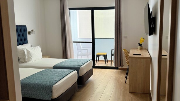A hotel room with two beds, a desk, a chair, a TV, and a balcony with a view. - Hotel Blue Sky (Tarrafal)