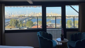 Habitación de hotel con amplias ventanas que ofrecen vistas a un paisaje urbano, un río y un puente. Hay dos sillas azules y una pequeña mesa con una planta en maceta.