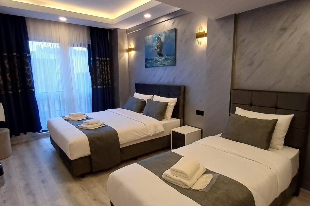 A modern hotel room with two beds, a large window with curtains, a painting on the wall, and a marble-textured wall.