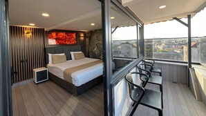 Un dormitorio moderno con una cama grande, una mesita de noche y una silla. Se aprecia vista de la ciudad a través de amplias ventanas.