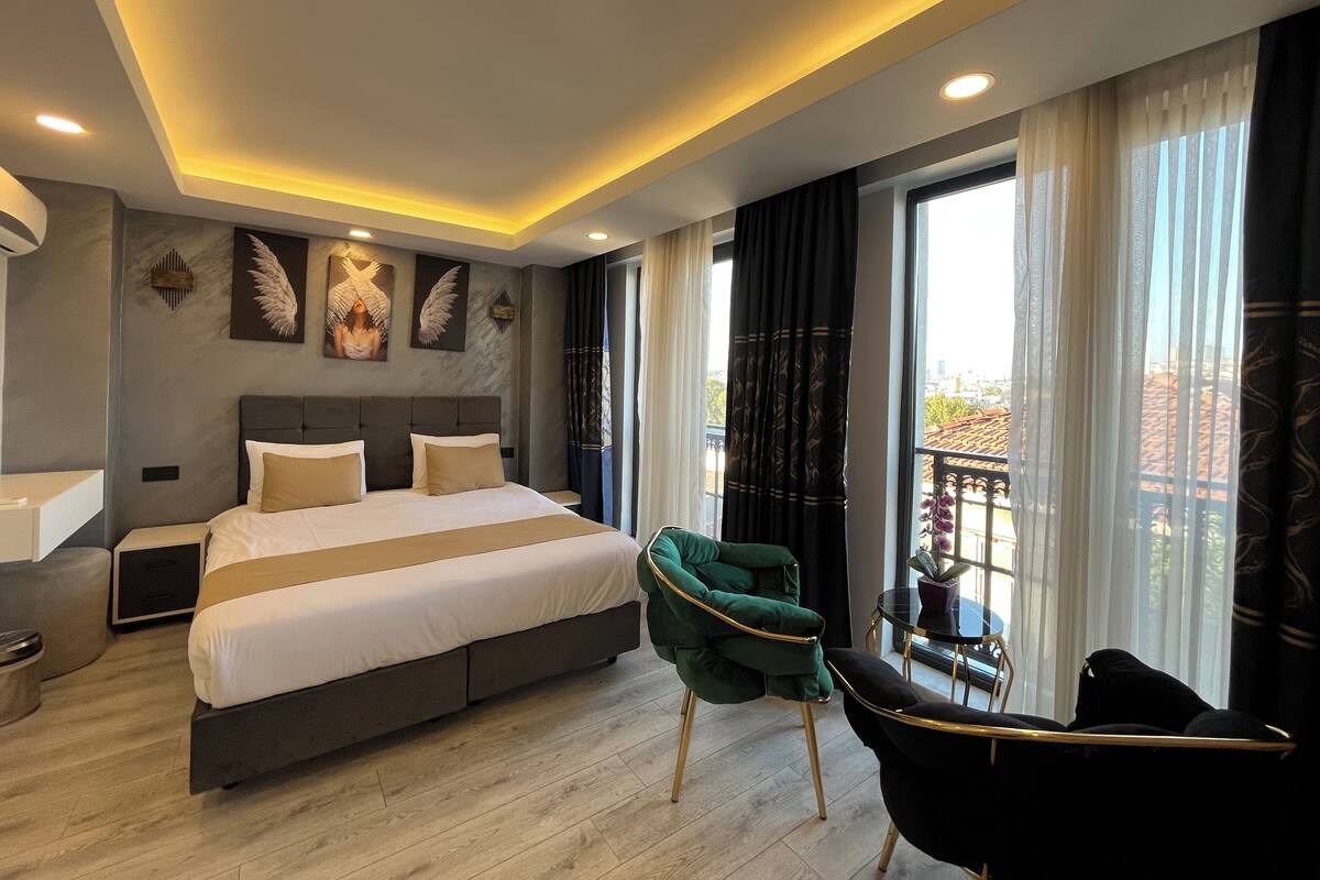 A modern hotel room with a large bed, a green armchair, a black chair, and a balcony with a view.