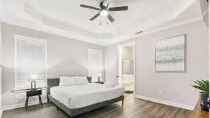 Ein Schlafzimmer mit Bett, Nachttischen, Deckenventilator, Fenster mit Jalousien, einer Tür zu einem Badezimmer und einem gerahmten Bild an der Wand.