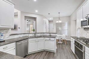 Une cuisine moderne avec des meubles blancs, un lave-vaisselle encastré, une cuisinière en acier inoxydable et un coin repas comprenant une table et des chaises blanches.