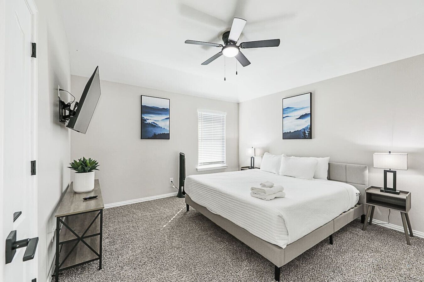 Une chambre à coucher moderne avec un lit, un ventilateur de plafond, un téléviseur fixé au mur et deux tableaux encadrés.