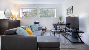 Una sala de estar moderna con un sofá seccional gris, una mesa de centro, un televisor y un ventanal con vista a árboles.