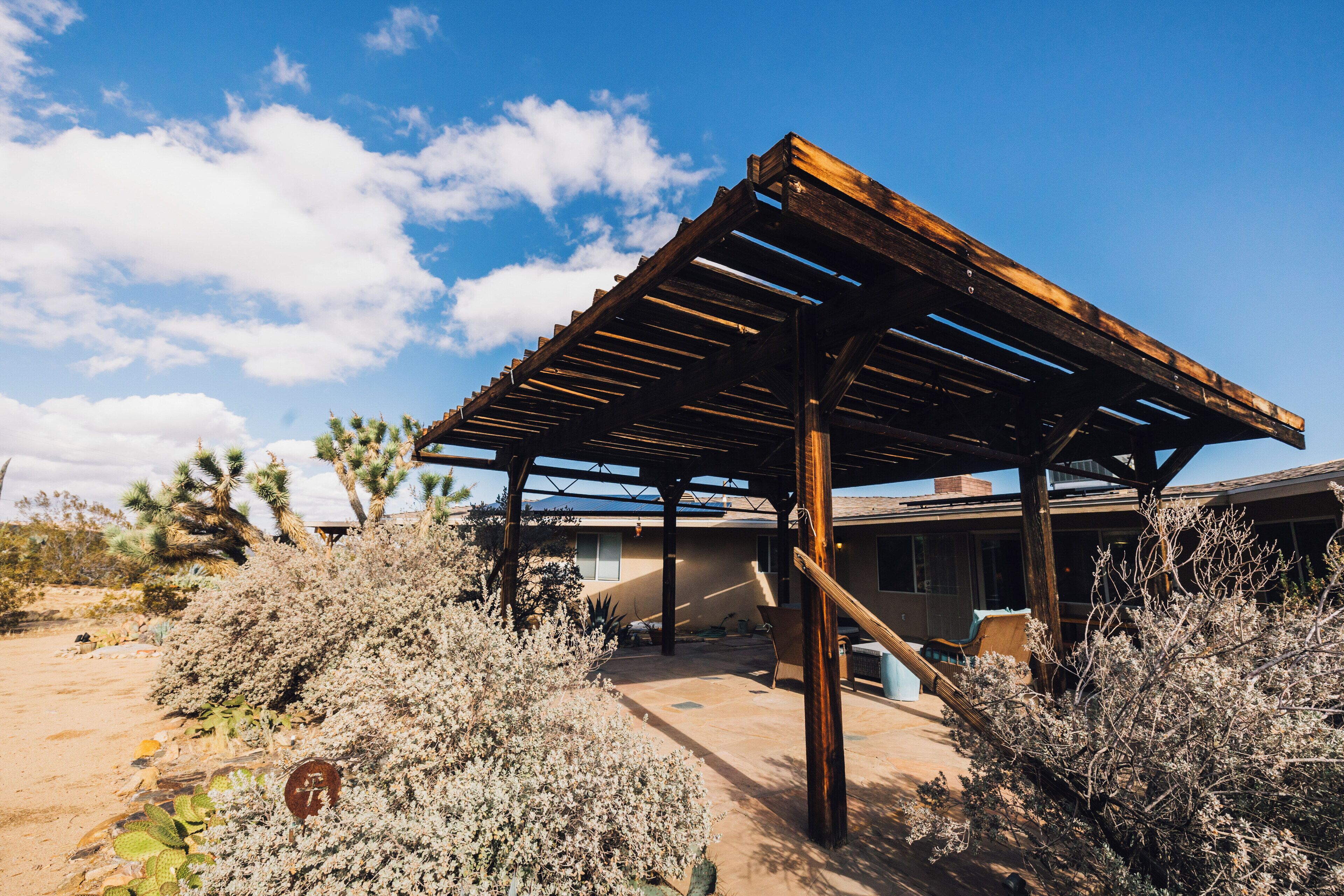 A covered outdoor area with a wooden pergola, surrounded by desert vegetation and a building in the background.