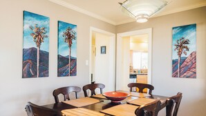 A dining area with a wooden table, chairs, and a red bowl centerpiece. There are paintings of palm trees on the walls. - 5 Miles from Park entrance Charming 2-bedroom house in enchanting Joshua Tree (Joshua Tree)