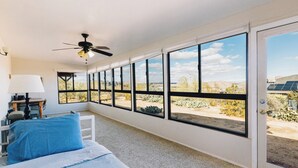 A bedroom with a bed, a desk, and a chair. There are large windows with a view of the desert. - 5 Miles from Park entrance Charming 2-bedroom house in enchanting Joshua Tree (Joshua Tree)
