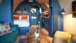 A cozy room with a bed, a green sofa, a wooden table with a bottle and glasses, and a small table lamp. - Luxury Rural Cabin with Wood Fired Hot Tub (Leven)