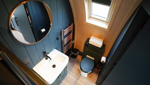 Ein kleines Badezimmer mit rundem Spiegel, Waschbecken, Toilette und kleinem Fenster.