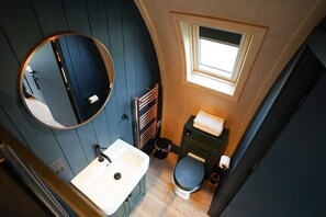A compact bathroom with a round mirror, a sink, a toilet, and a small window. - Luxury Rural Cabin with Wood Fired Hot Tub (Leven)
