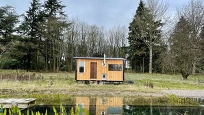 Eine kleine Holzhütte mit Tür und Fenstern, neben einem ruhigen Teich mit Spiegelungen, umgeben von Bäumen und Gras.