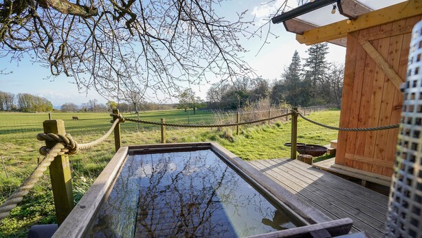 Eine Holzterrasse mit Seilgeländer, einem Holzbau und freiem Blick auf ein saftiges Grasfeld.