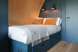 A bed with a white mattress and orange pillows, a blue headboard, and a small bedside table with a drawer.