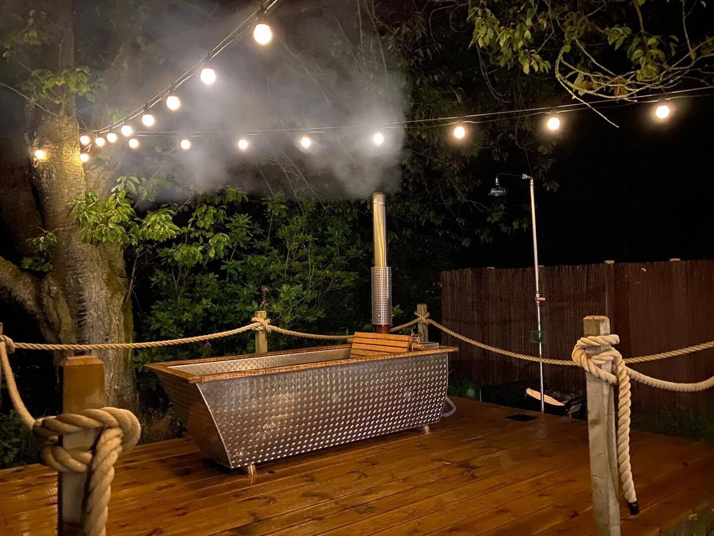 An outdoor wooden deck with a metal bench, surrounded by ropes and string lights.