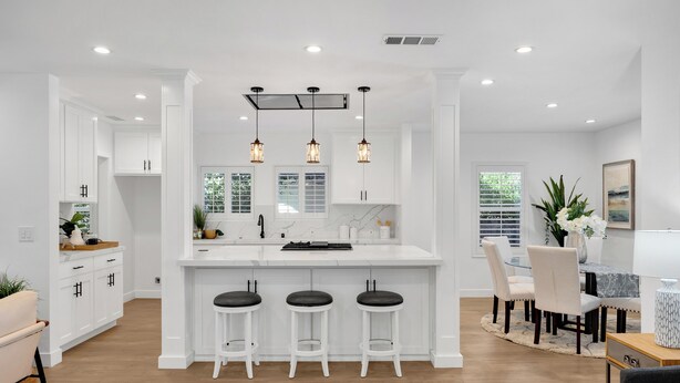 Une cuisine moderne avec des meubles blancs, un îlot central doté de tabourets, un éclairage suspendu et un coin repas avec une table et des chaises.