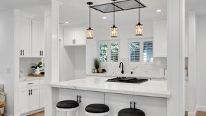 Une cuisine moderne avec des meubles blancs, un grand îlot central doté de tabourets, un éclairage suspendu et une vue sur la verdure à l’extérieur.