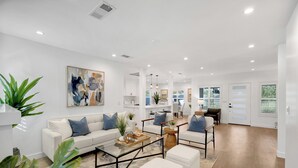 Un salon moderne avec un canapé blanc, une table basse et un pouf, un coin repas avec des chaises, et une cuisine en arrière-plan.