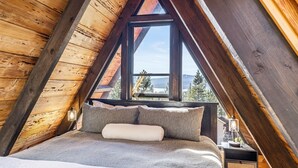 Ein gemütliches Schlafzimmer unter dem Dach mit holzverkleideter Schräge, einem großen Fenster mit Blick ins Freie und einem Nachttisch mit Lampe.