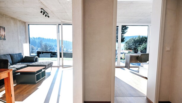 Amplia sala de estar con sofá, mesa de centro y una silla de madera. Un gran ventanal ofrece vistas al exterior.