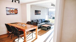 Un espacio de estar moderno con una mesa de comedor de madera, sillas negras, un sofá gris y una mesa de centro. Hay dos cuadros en las paredes y un ventanal que ofrece vistas al exterior.