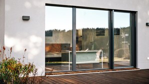 Un edificio moderno con amplios ventanales de vidrio, una terraza de madera y vistas a una piscina y árboles.