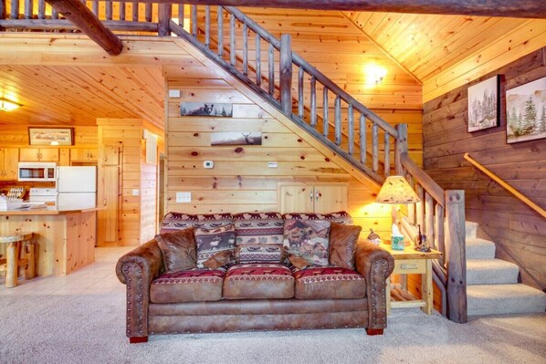A cozy cabin interior with a wooden staircase, a sofa with patterned cushions, a kitchen area, and a dining table.
