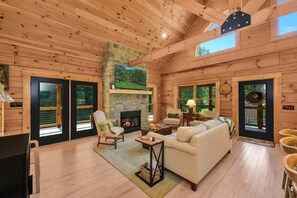 A spacious living room with a stone fireplace, wooden walls, and large windows.