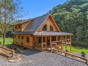 A wooden cabin with a porch and a deck, surrounded by a forest and a lake.