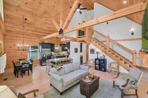 A spacious living area with a sofa, chairs, a dining table, and a kitchen in the background. A wooden staircase leads to an upper level.