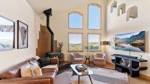 A cozy living room with a fireplace, comfortable seating, large windows, and a flat-screen TV.