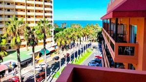 Un hôtel de grande hauteur offrant une vue sur l’océan, avec des palmiers bordant la rue et un ciel bleu dégagé.