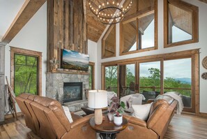 A spacious living room with leather sofas, a fireplace, a large chandelier, and a scenic view through large windows. - Luxury Eagles Nest Lodge: Hot Tub, Game Loft, Fire Pit, & Year-Round Panoramic Views! (Elk Park)