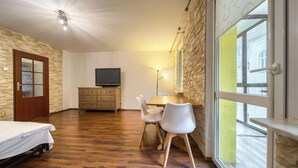Ein Zimmer mit Holzboden, einem Bett, einem Fernseher auf einer Holzkommode, einem Schreibtisch mit Stühlen und einem Fenster mit Blick auf Gebäude.
